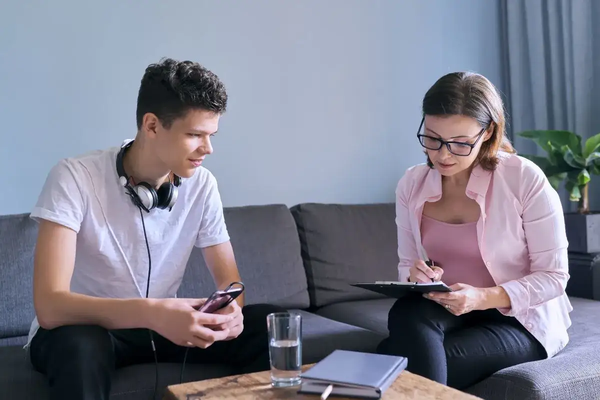 Psycholog młodzieży rozmawia z nastolatkiem, który trzyma telefon. Kobieta notuje na tablicy.