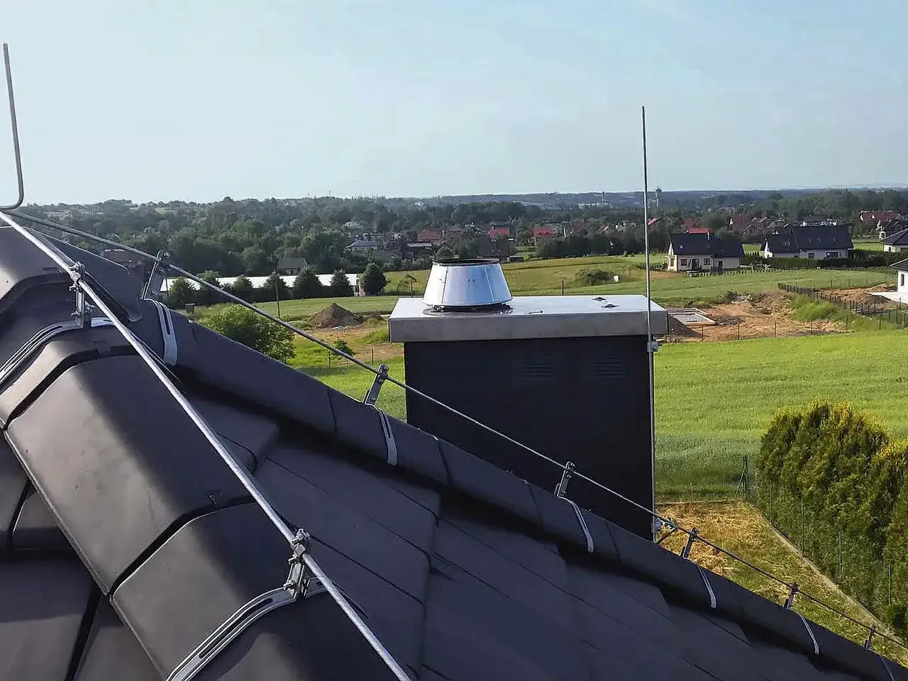 Nowy dach z czarnej dachówki, na którym zamontowano instalację odgromową. W tle zielone pola i domy.