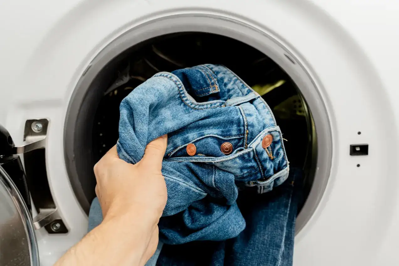 Ręka wkłada dżinsy do pralki. Czas na program denim w pralce, by odświeżyć ulubione spodnie.