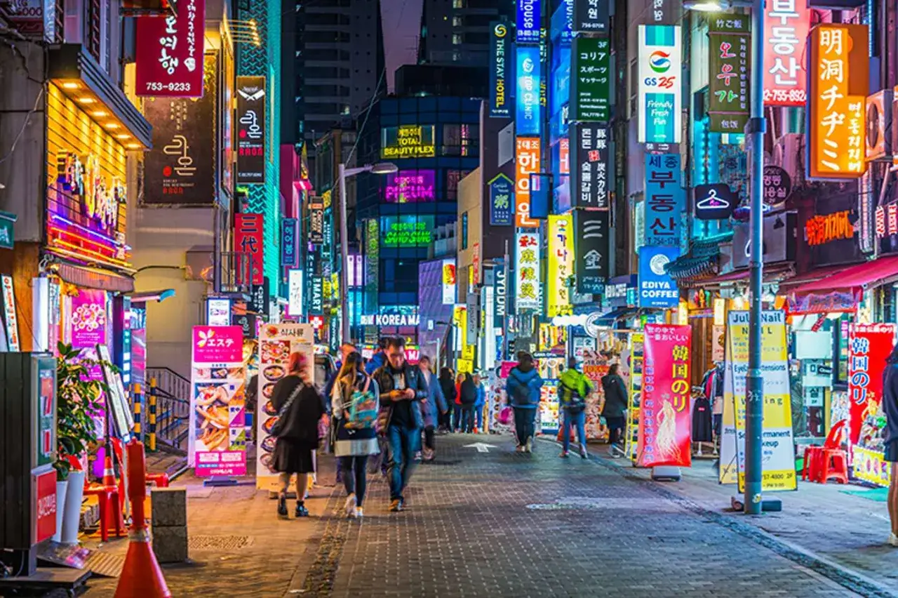 Tętniąca życiem ulica w **Gangnam** nocą, pełna neonowych szyldów i ludzi spacerujących po chodniku.