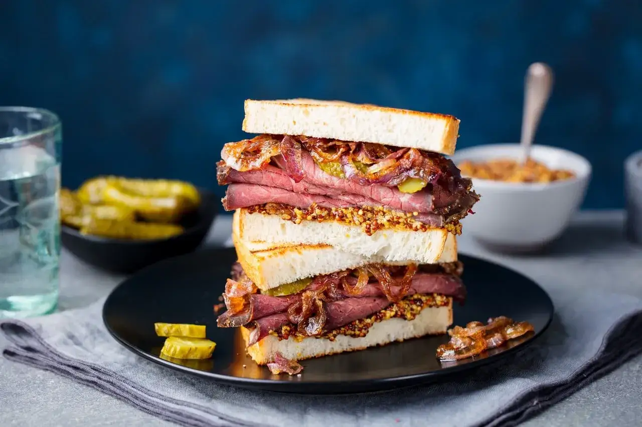 Un delicioso sándwich de pastrami como se come, con pan tostado, mostaza, pepinillos y cebolla caramelizada.
