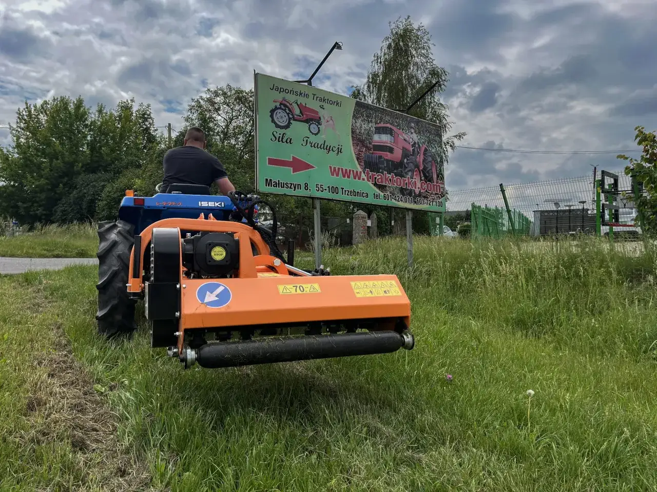 Mężczyzna za kierownicą traktorka Iseki z kosiarką tylno boczną, pracującą na tle reklamy 