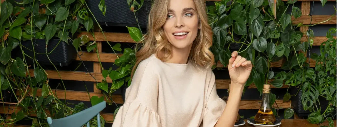 Uśmiechnięta blondynka w beżowej bluzce, otoczona bujną zielenią. Zastanawia się, jaki kolor pasuje do beżu, a zieleń wokół dodaje jej spokoju.