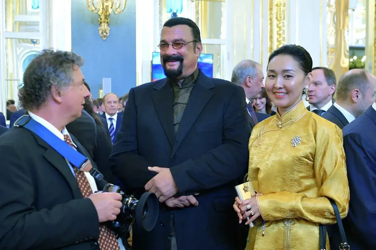 Steven Seagal z uśmiechem pozuje do zdjęcia z kobietą w żółtej sukni. W tle widać innych gości, w tym mężczyznę z aparatem, który może być ojcem Stevena Seagala, lub po prostu jego fanem.