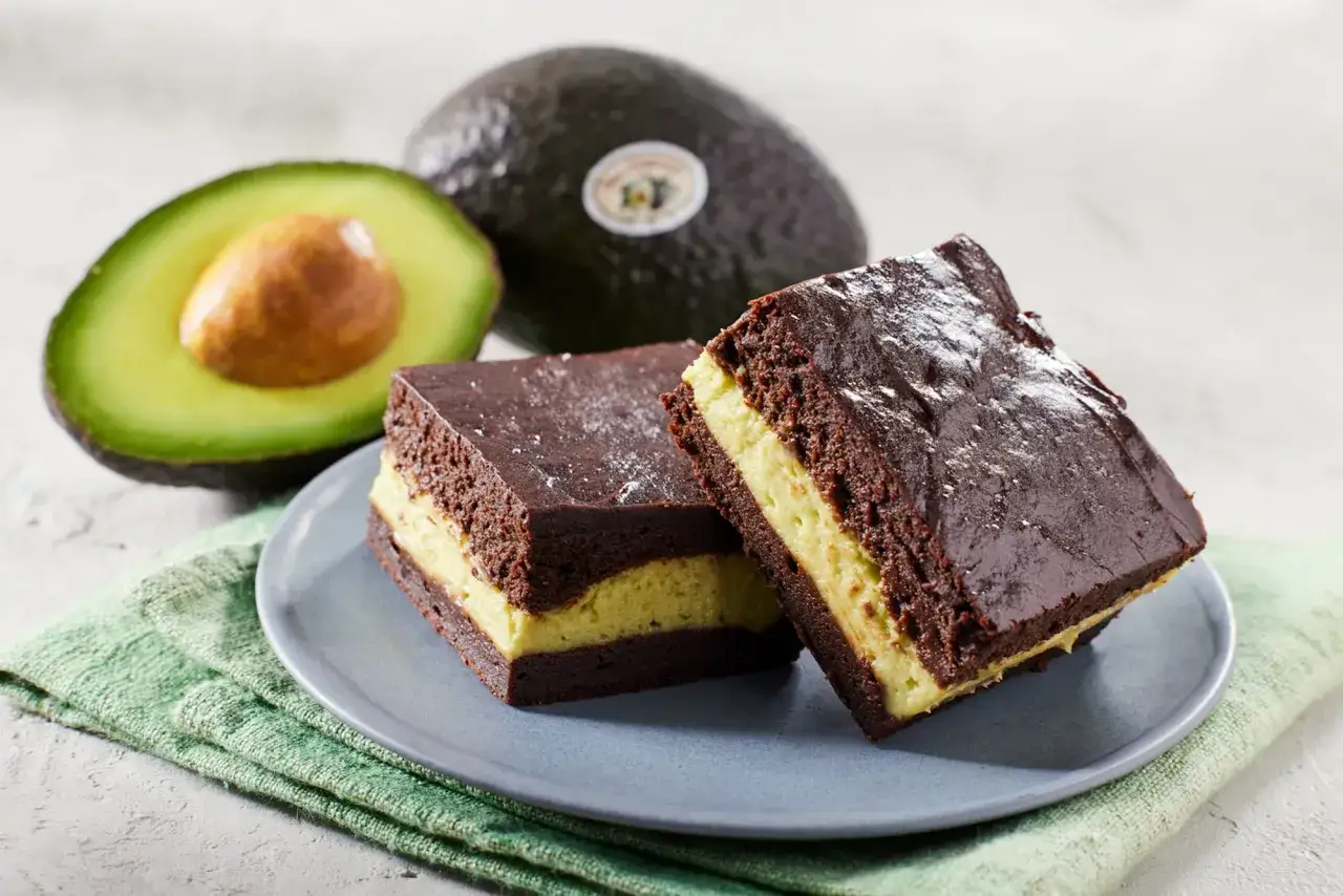 Dwa pyszne brownie z awokado, posypane cukrem pudrem, leżą na talerzyku obok świeżych awokado.