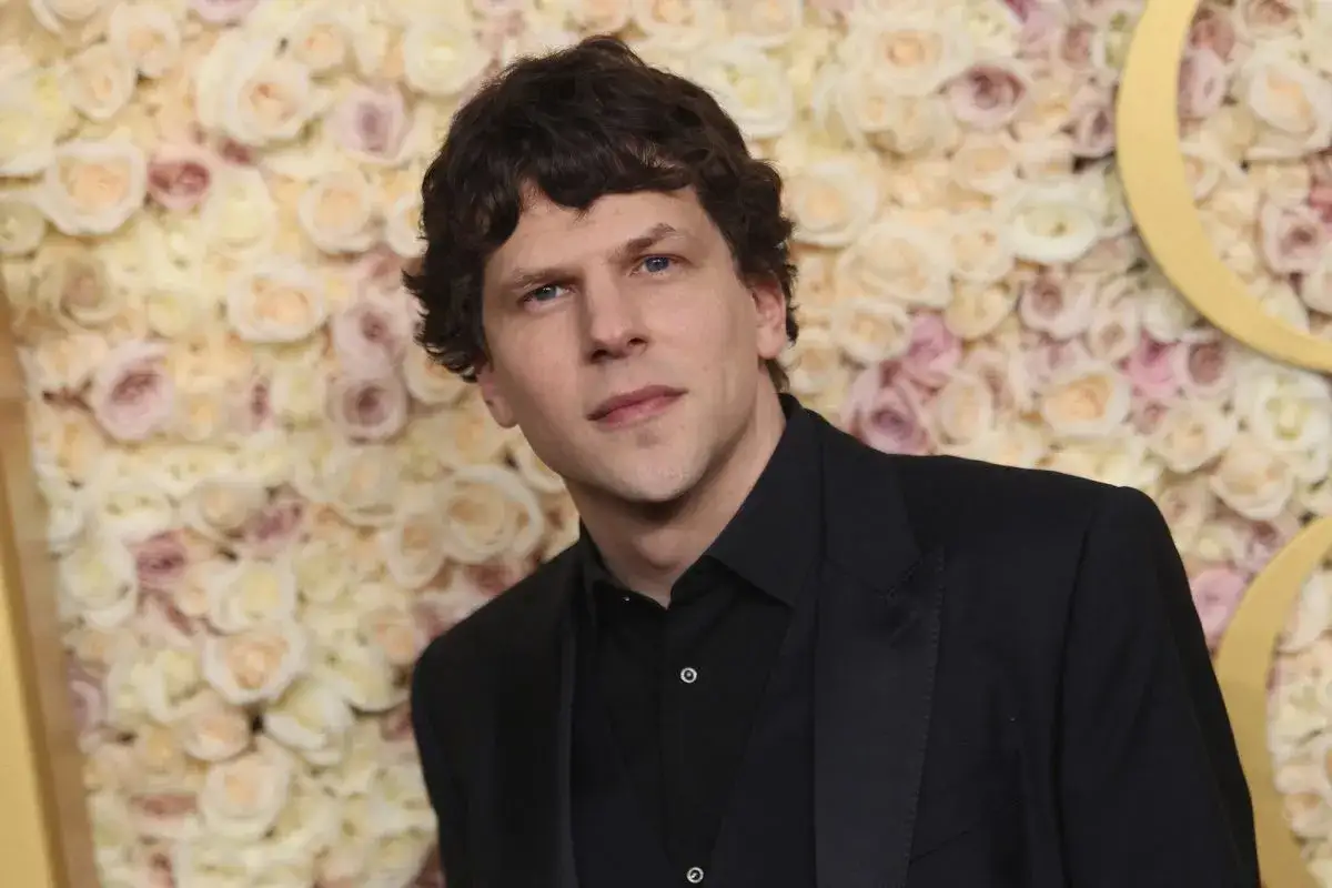 Jesse Eisenberg w czarnym smokingu na tle ściany z różowych i kremowych kwiatów.