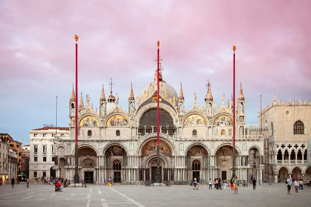 La maestosa basilica di San Marco a Venezia, con le sue cupole dorate e mosaici scintillanti, si staglia contro un cielo rosa al tramonto.