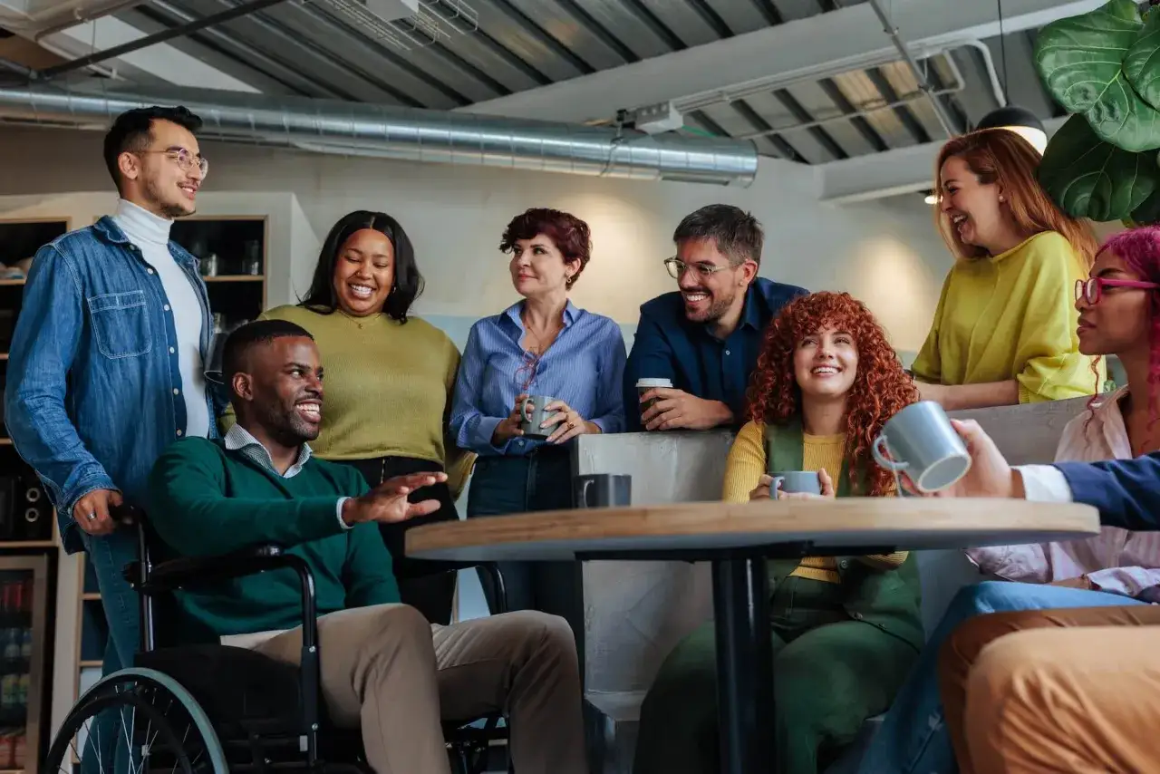 A diverse group of colleagues, including a man in a wheelchair, share a laugh. They seem to be in a casual meeting, perhaps with an **internal coach** guiding their discussion.