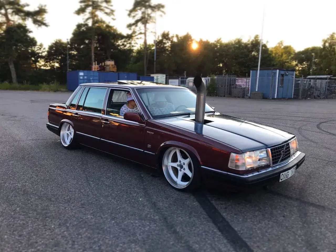 En mörkröd Volvo 760 D24 med en vertikal pipa som sticker upp från motorhuven. Bilen har vita fälgar och är parkerad utomhus.