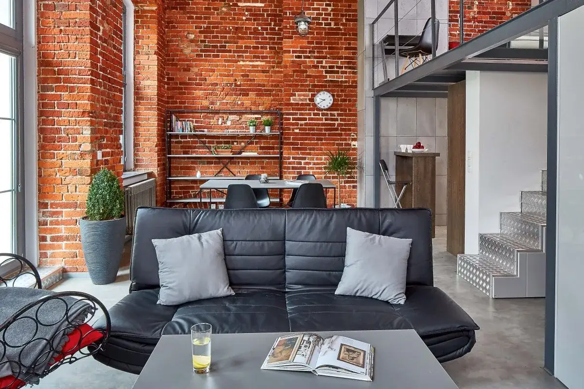 Nowoczesny salon w stylu loftowym z ceglaną ścianą, czarną kanapą i metalowymi schodami na antresolę.