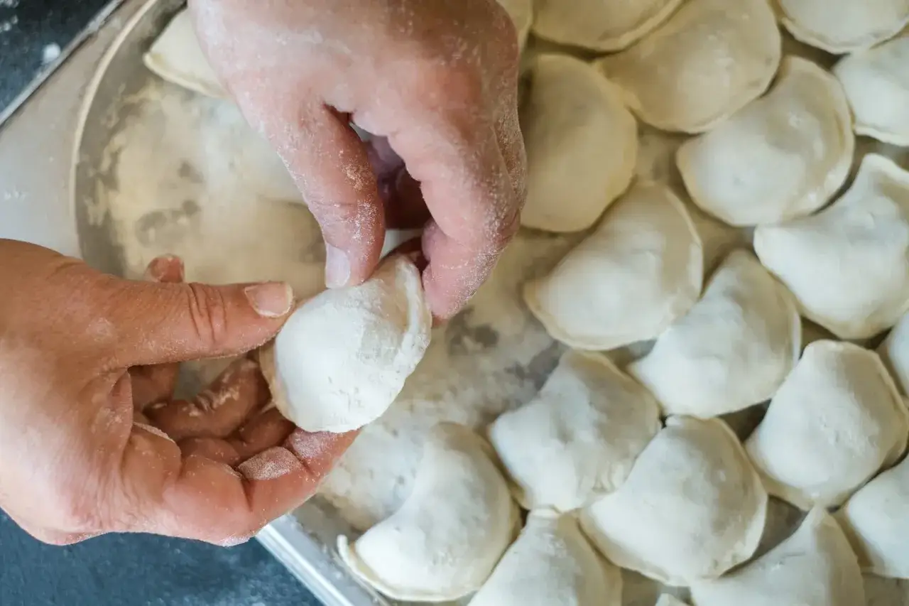 Dłonie formują ciasto na pierogi, które leżą obok siebie na blacie.
