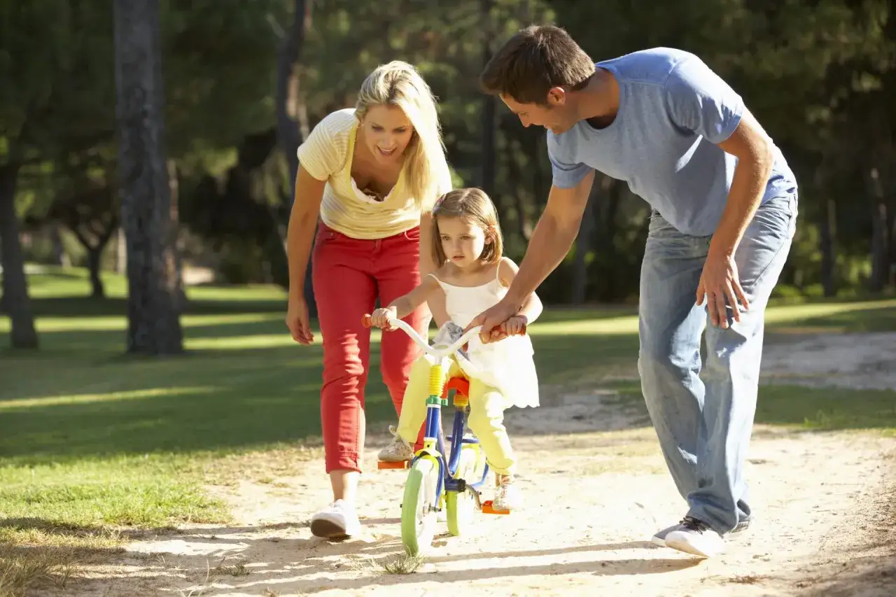 Genitori aiutano bambina a imparare ad andare in bici, un modo divertente per stimolare il linguaggio nei bambini con nuove parole ed esperienze.