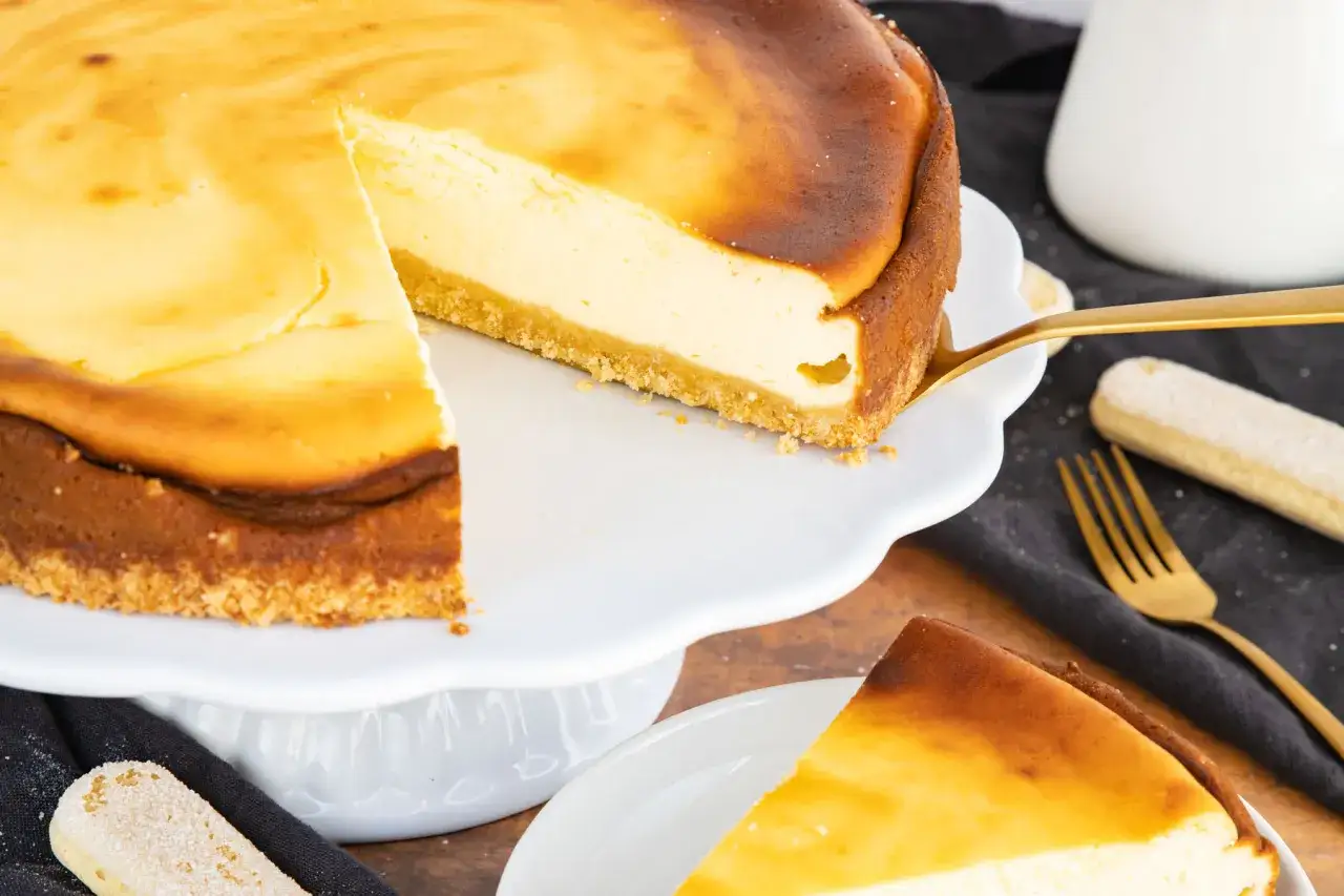 Cremiger cheesecake mit keksboden, eine Scheibe wird mit einem goldenen Tortenheber herausgenommen.