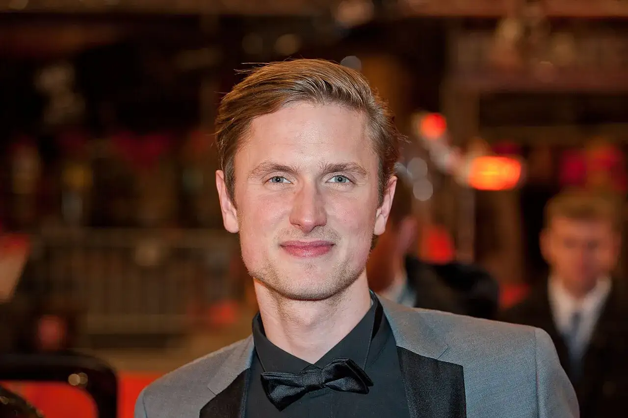 Mikkel Boe Følsgaard, en skådespelare med blont hår och blå ögon, bär en grå kavaj och en svart fluga.