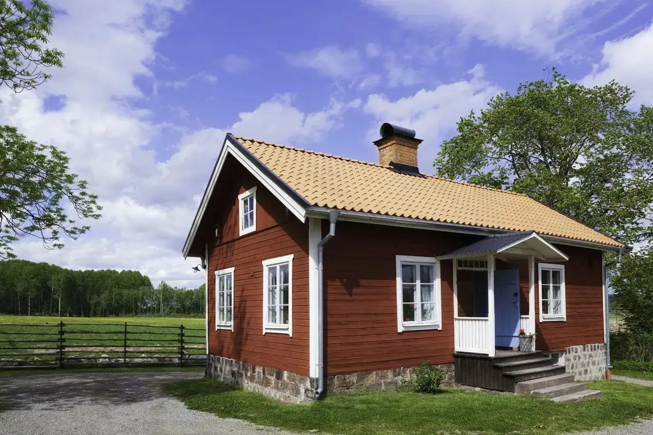 Rött trähus med tegeltak och skorsten. Planer på att bygga ut gammalt hus i lantlig miljö med äng och skog i bakgrunden.