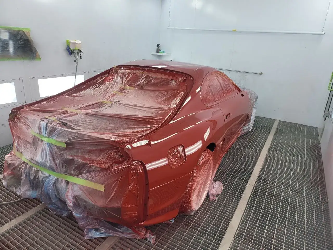 Röd bil i en lackbox, redo för att lacka om bilen. Skyddsplast täcker större delen av fordonet.