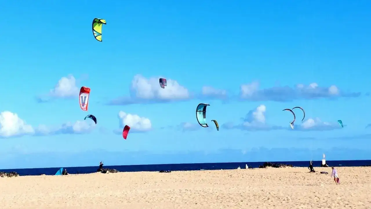 Fuerteventura pogoda styczeń: kolorowe latawce na błękitnym niebie nad piaszczystą plażą i oceanem. Idealne warunki do kitesurfingu.