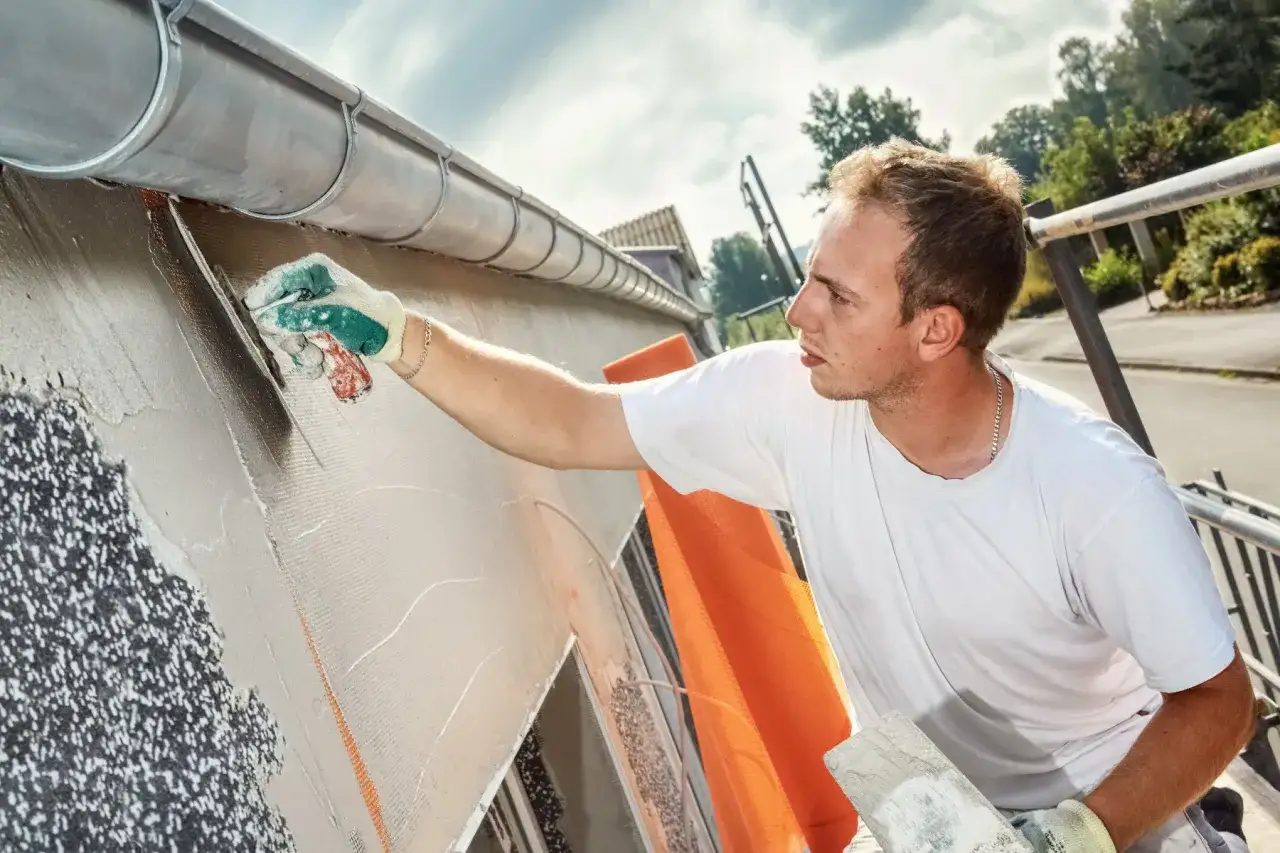 Un ouvrier applique un ragréage sur un mur extérieur, utilisant une truelle pour lisser le mortier sur une armature.
