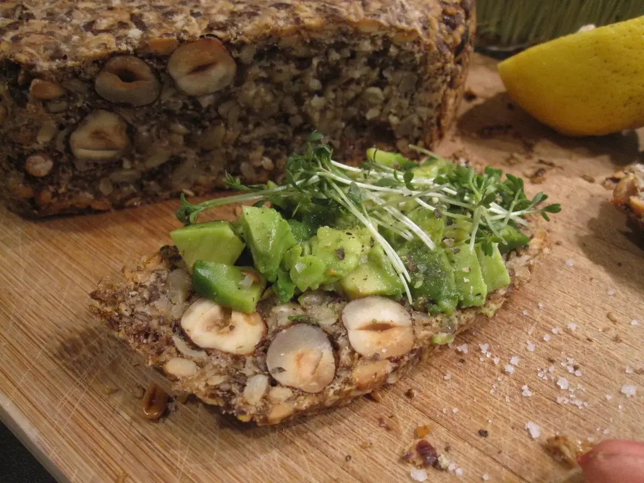 Ein Stück **flohsamenschalen brot** mit Avocado und Sprossen belegt, daneben ein ganzer Laib und eine Zitronenspalte.