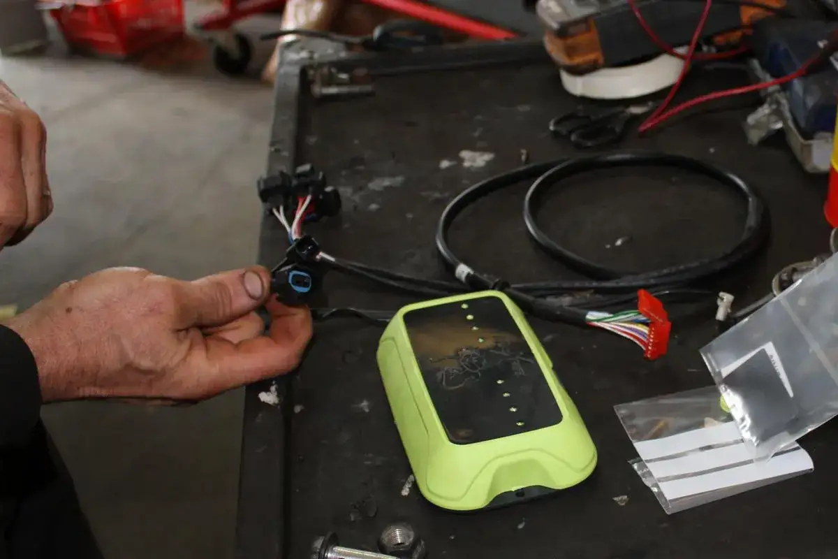 Main d'un m&eacute;canicien tenant un connecteur pour l'installation d'un bo&icirc;tier &eacute;thanol. Le bo&icirc;tier vert et les c&acirc;bles sont visibles sur un &eacute;tabli.