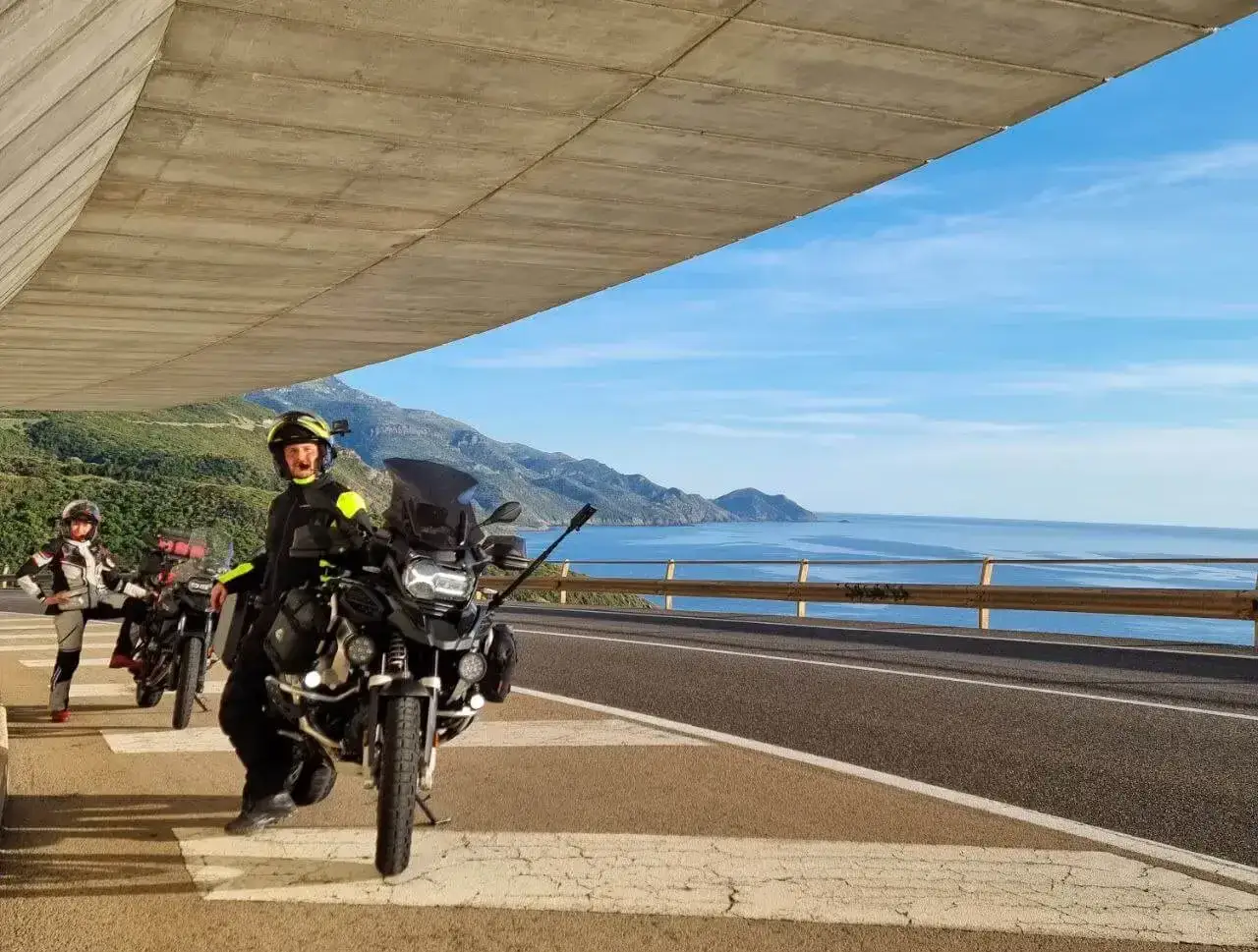 Due motociclisti si fermano per una foto panoramica sul passo Bidderdi, con il mare blu e le montagne sullo sfondo.