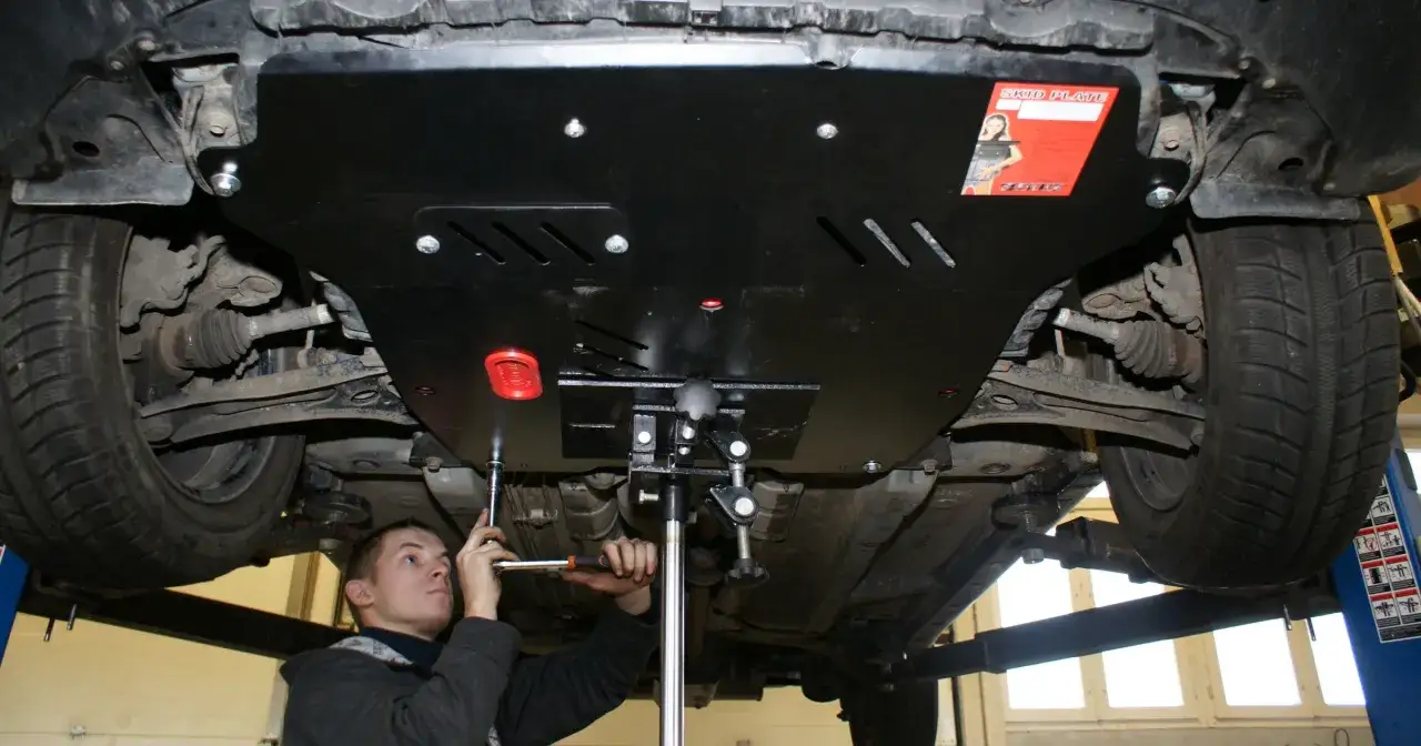 Młody mechanik dokręca śruby, aby jak zamocować luźną osłonę pod silnikiem do zderzaka.