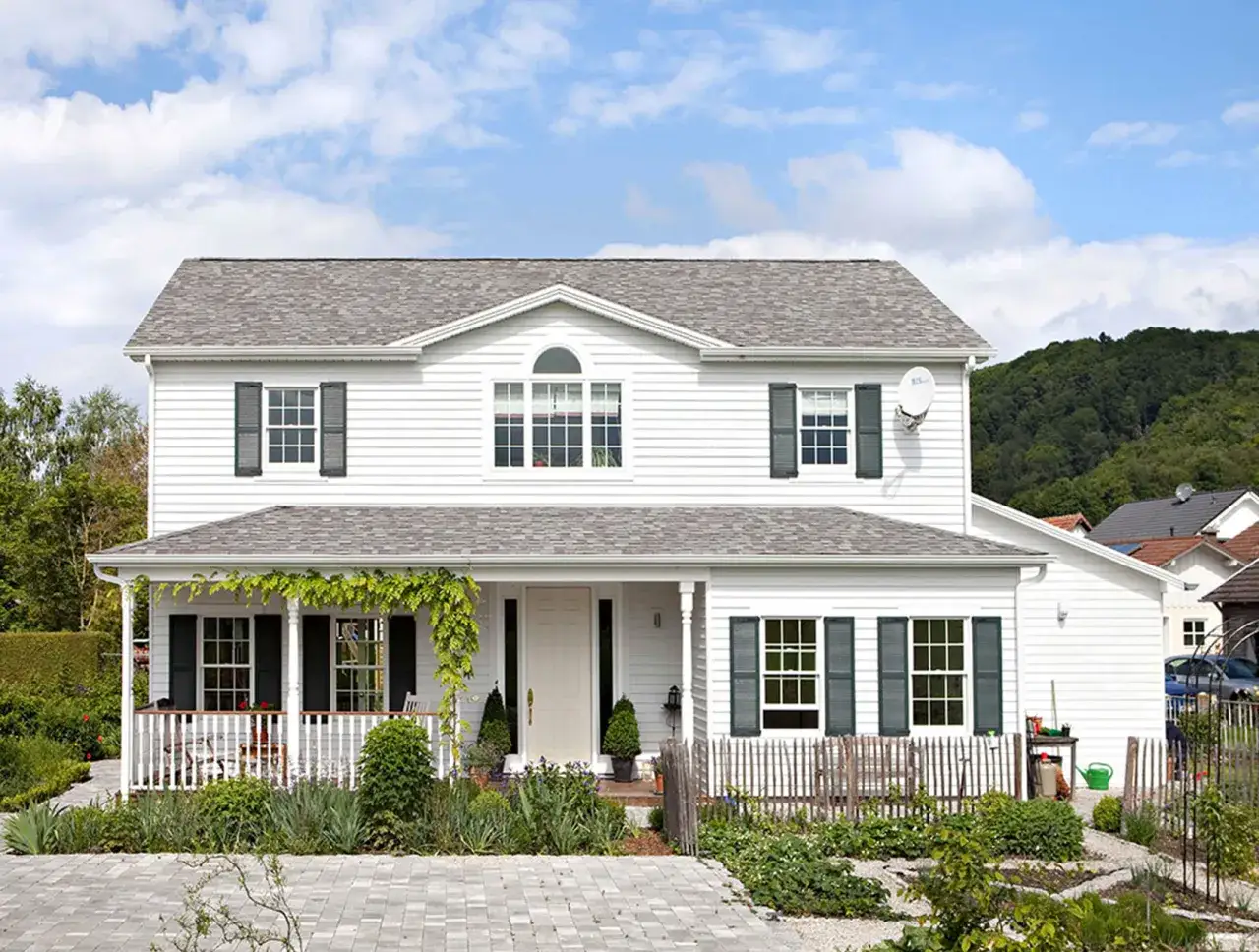 Ein wei&szlig;es Haus im amerikanischen Stil mit Veranda, gr&uuml;nen Fensterl&auml;den und einem gepflegten Garten.