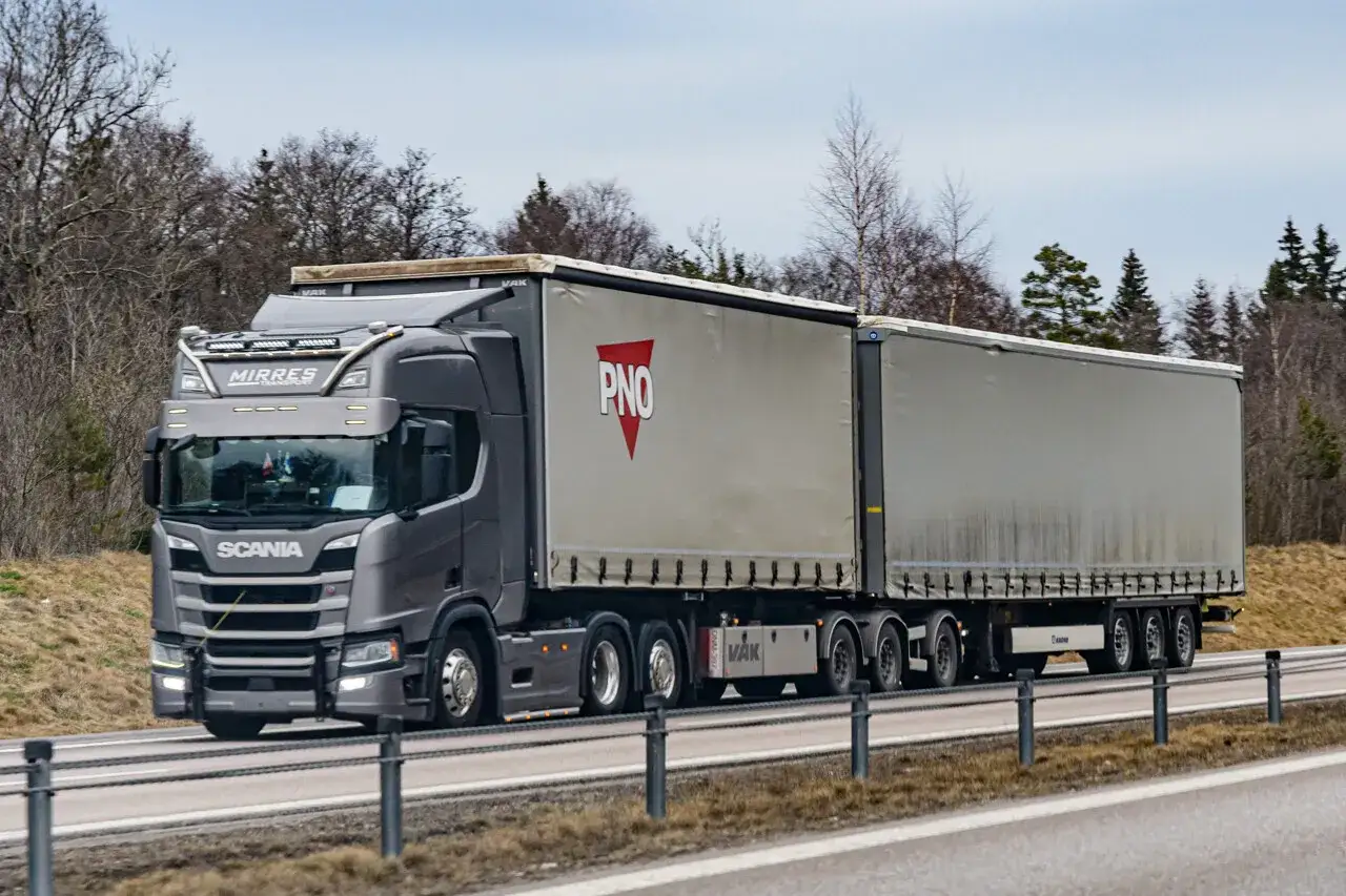 Un camion Scania gris, un camion 60 tonnes, roule sur une route bordée d'arbres. La remorque porte le logo PNO.