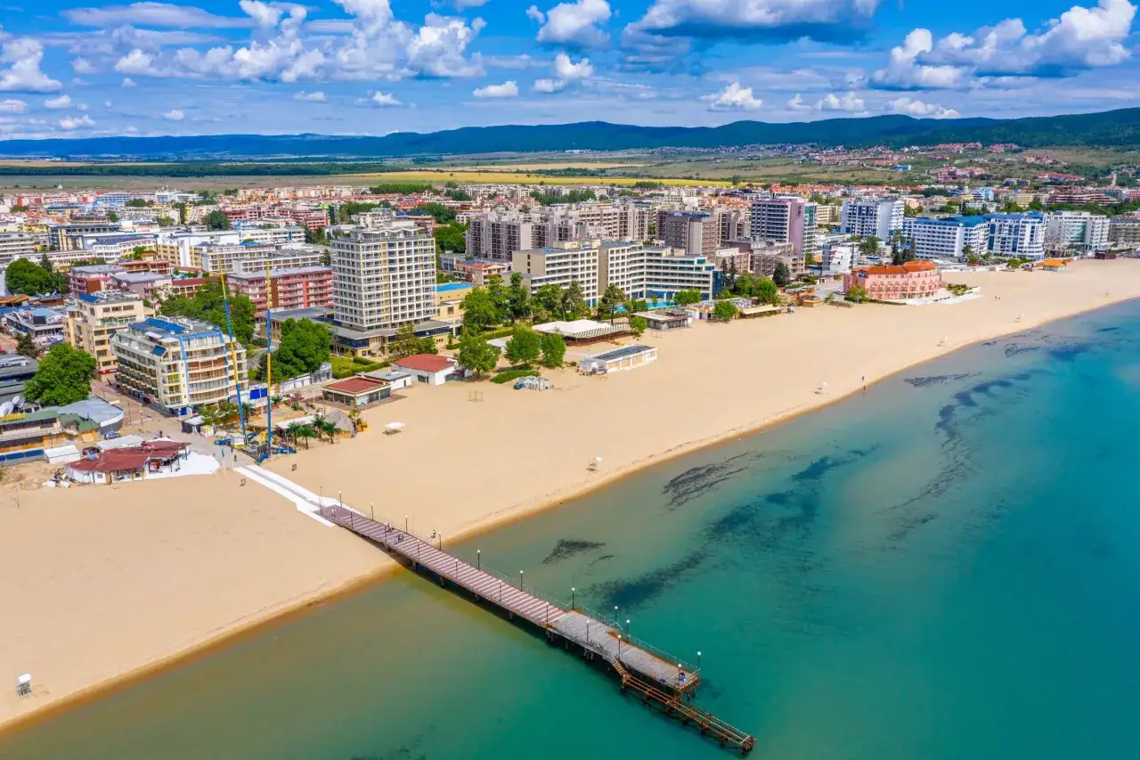 Słoneczny Brzeg: szeroka, piaszczysta plaża z długim molo, turkusowe morze i nadmorskie miasto z hotelami.
