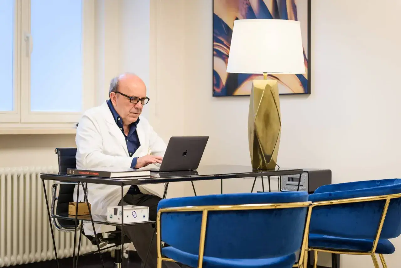 Lekarz w białym kitlu pracuje przy laptopie w gabinecie z niebieskimi fotelami.