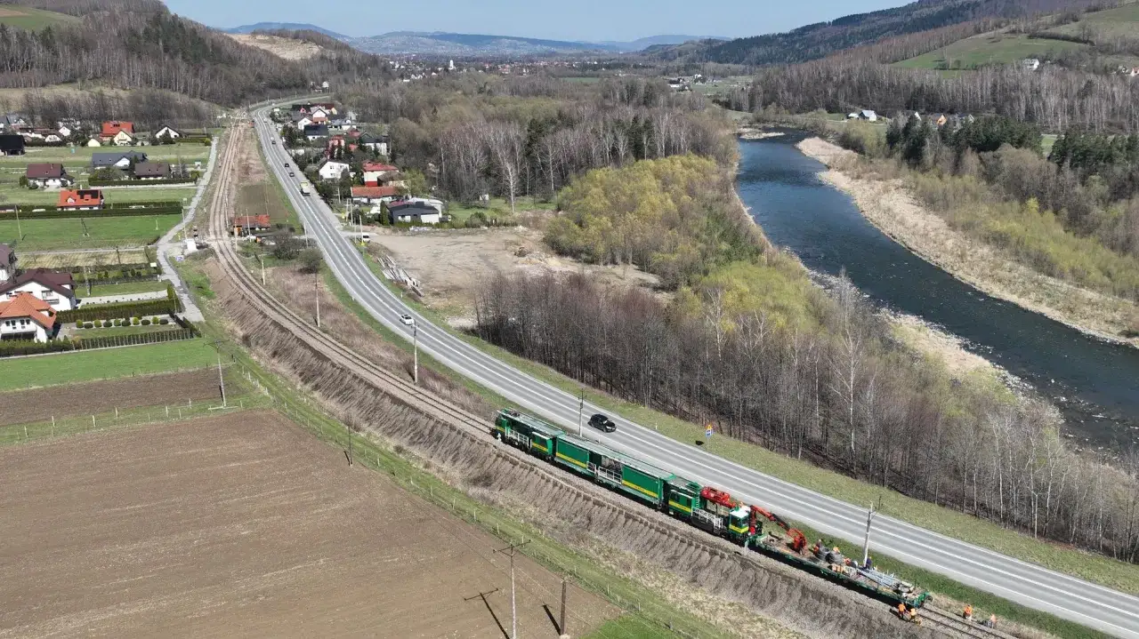 Zielony pociąg z żurawiem jedzie po torach w pobliżu drogi i rzeki. To może być pociąg techniczny w okolicach Nowego Sącza.