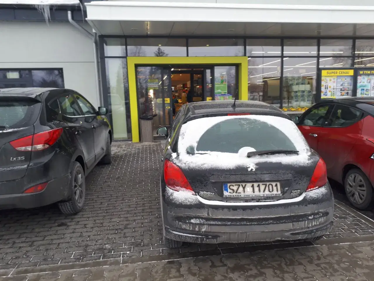 Samochody pod sklepem, jeden z nich ma tablicę rejestracyjną SZY 11701.