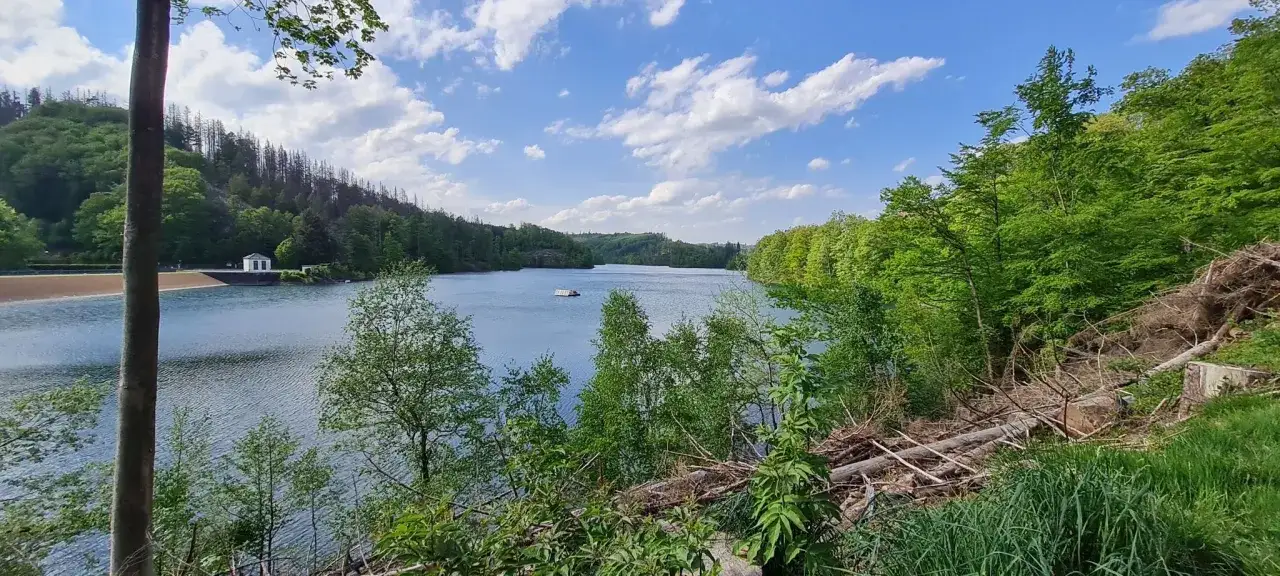 Idyllische Szene am genkeltalsperre rundweg: blauer Himmel mit Wolken, gr&uuml;ner Wald und ein ruhiger See mit einem kleinen Boot.