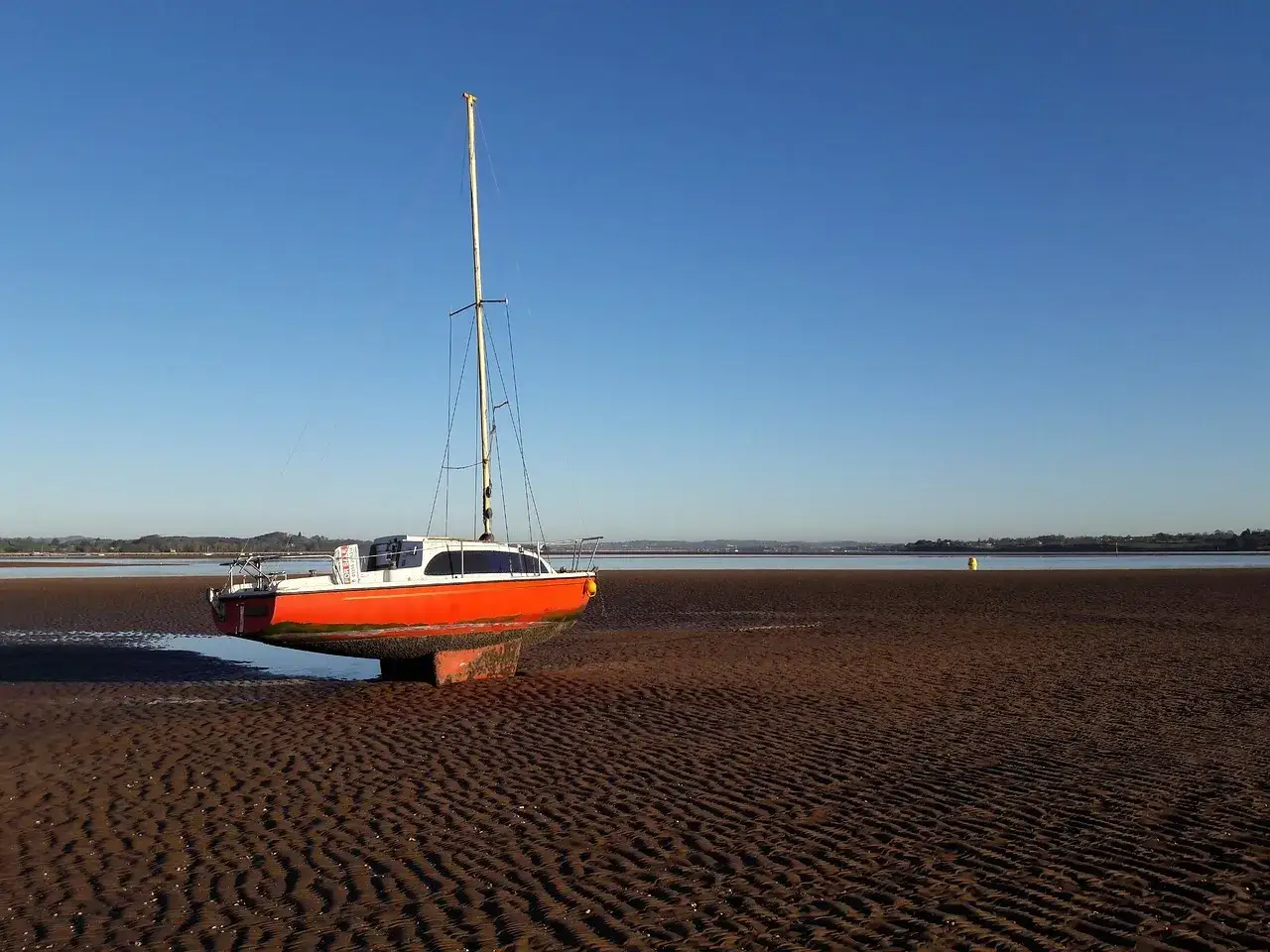 Un voilier anglais, échoué sur une plage de sable fin aux reflets orangés, attend la marée sous un ciel bleu limpide.