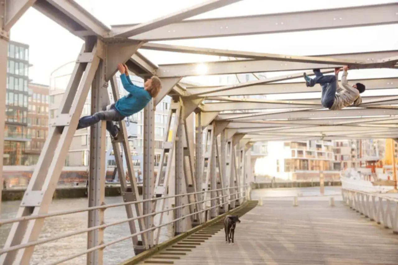 Zwei junge Männer klettern an einer Brücke in Hamburg. Ein Hund beobachtet sie.