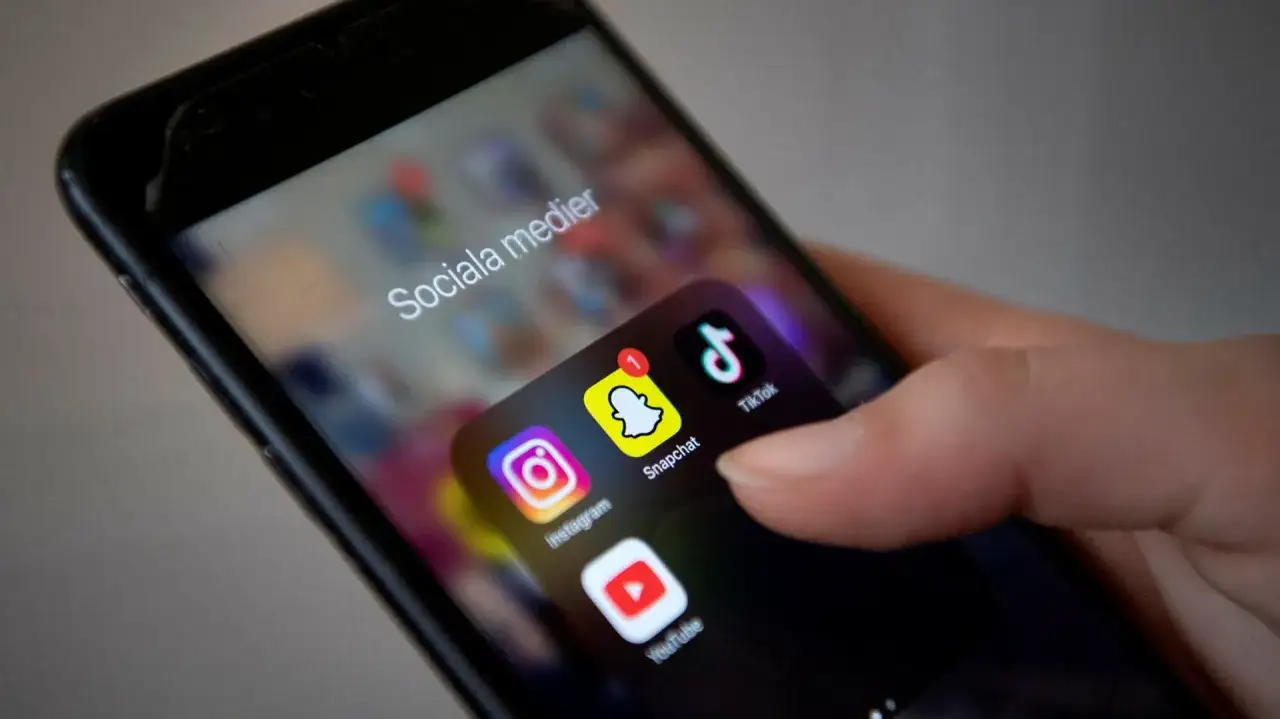 En hand h&aring;ller en mobiltelefon med ikoner f&ouml;r Instagram, Snapchat, TikTok och YouTube. Texten 