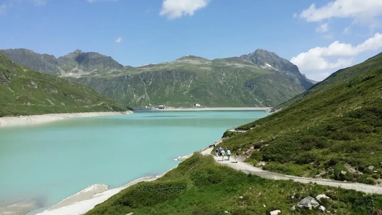 Wanderer genießen die Aussicht auf den türkisfarbenen Silvretta Stausee, umgeben von grünen Bergen unter blauem Himmel.