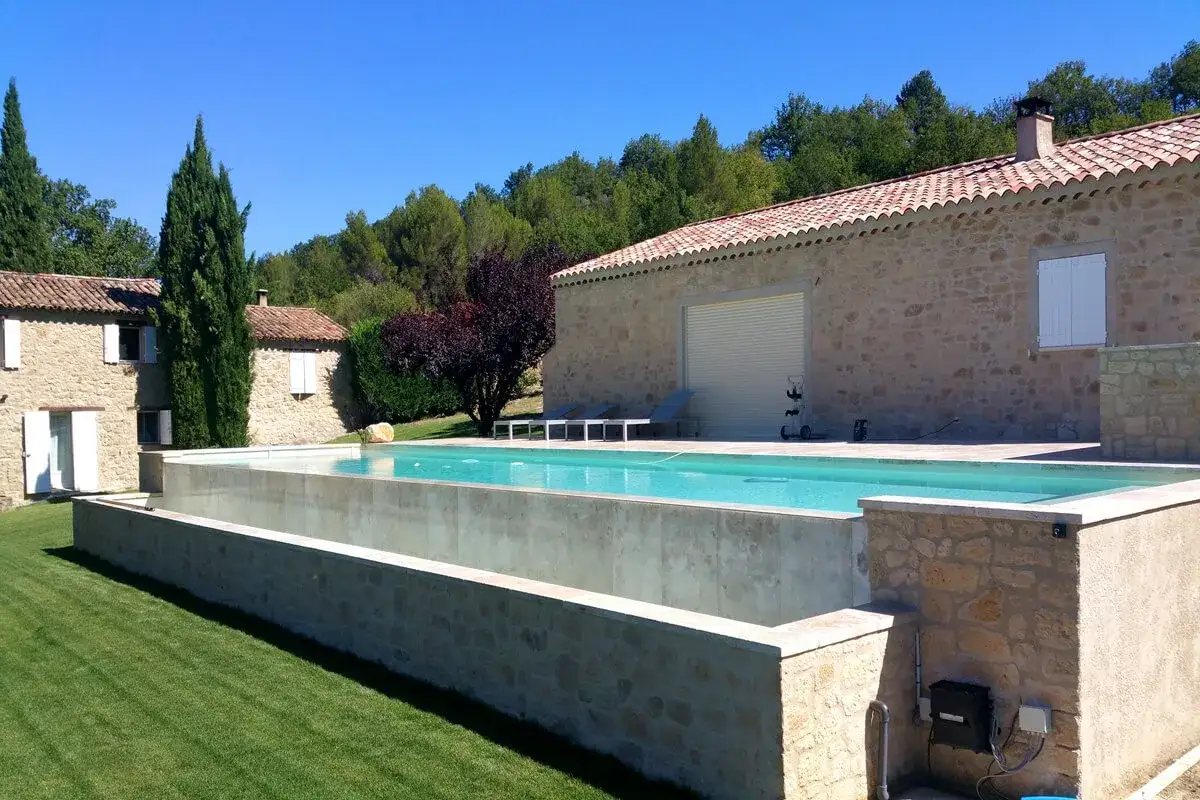 Elegantes piscinas elevadas de obra con revestimiento de cemento, rodeadas de c&eacute;sped verde y una casa de piedra bajo un cielo azul.