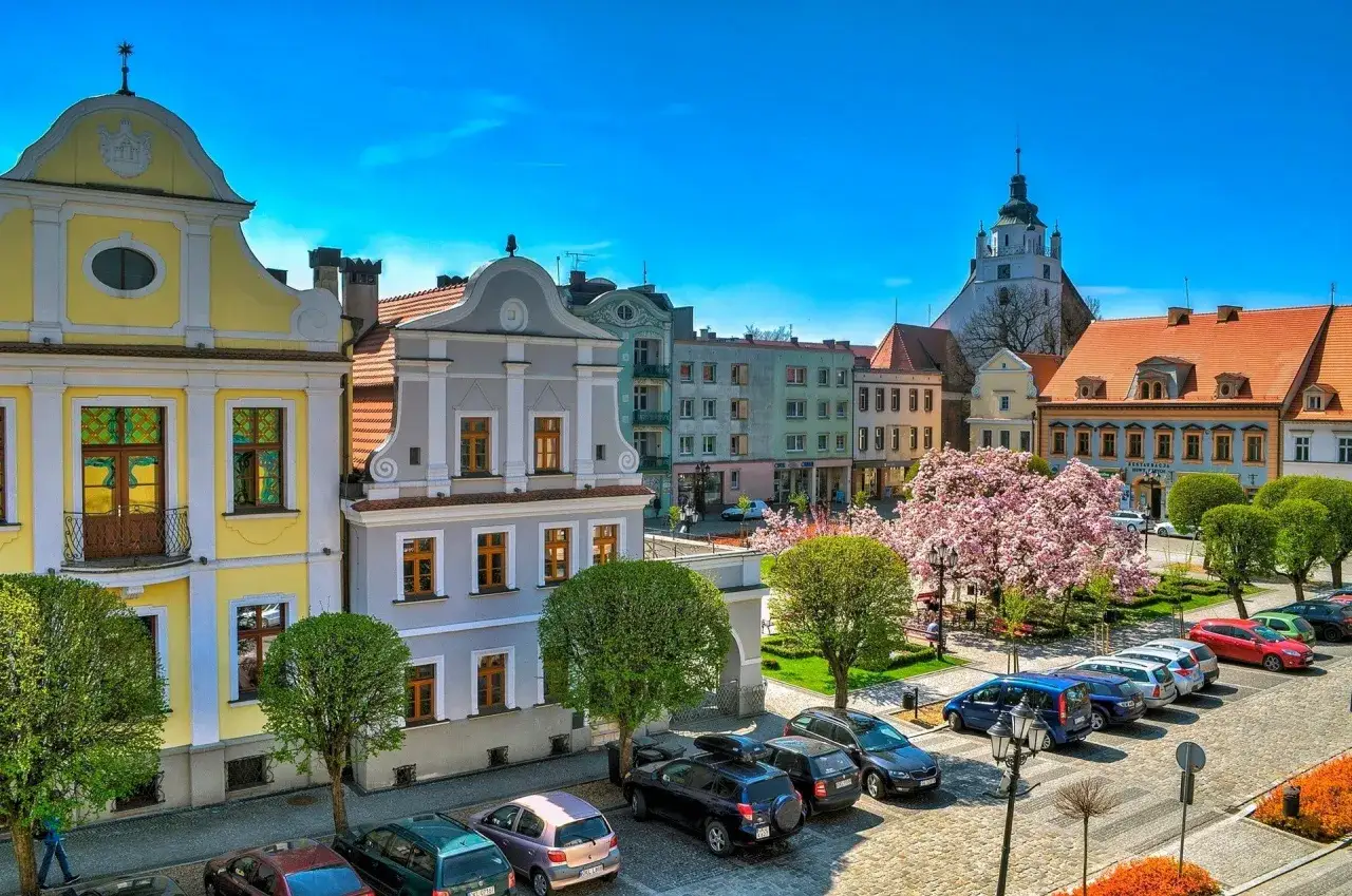 Urokliwy rynek w Kluczborku z kolorowymi kamienicami, kwitnącym drzewem i wieżą kościelną. Kluczbork atrakcje zachwycają!