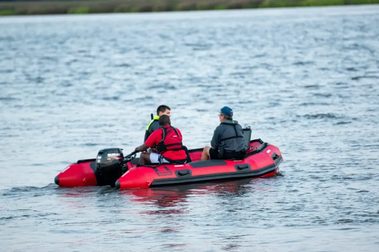 Rotes Schlauchboot mit drei Männern, die auf dem Wasser fahren. Einer steuert, die anderen schauen. Ein Mann trägt eine Rettungsweste. Das Boot scheint Wasser aufzunehmen, als ob es sinken würde.
