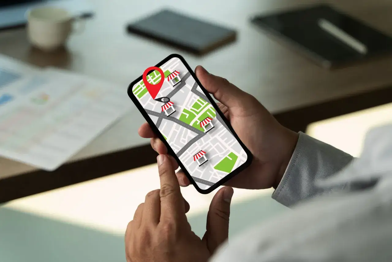 Dłoń trzyma smartfon z mapą i ikonami sklep&oacute;w. Czerwony pin wskazuje lokalizację. To reklama przypominająca o pobliskich punktach usługowych.