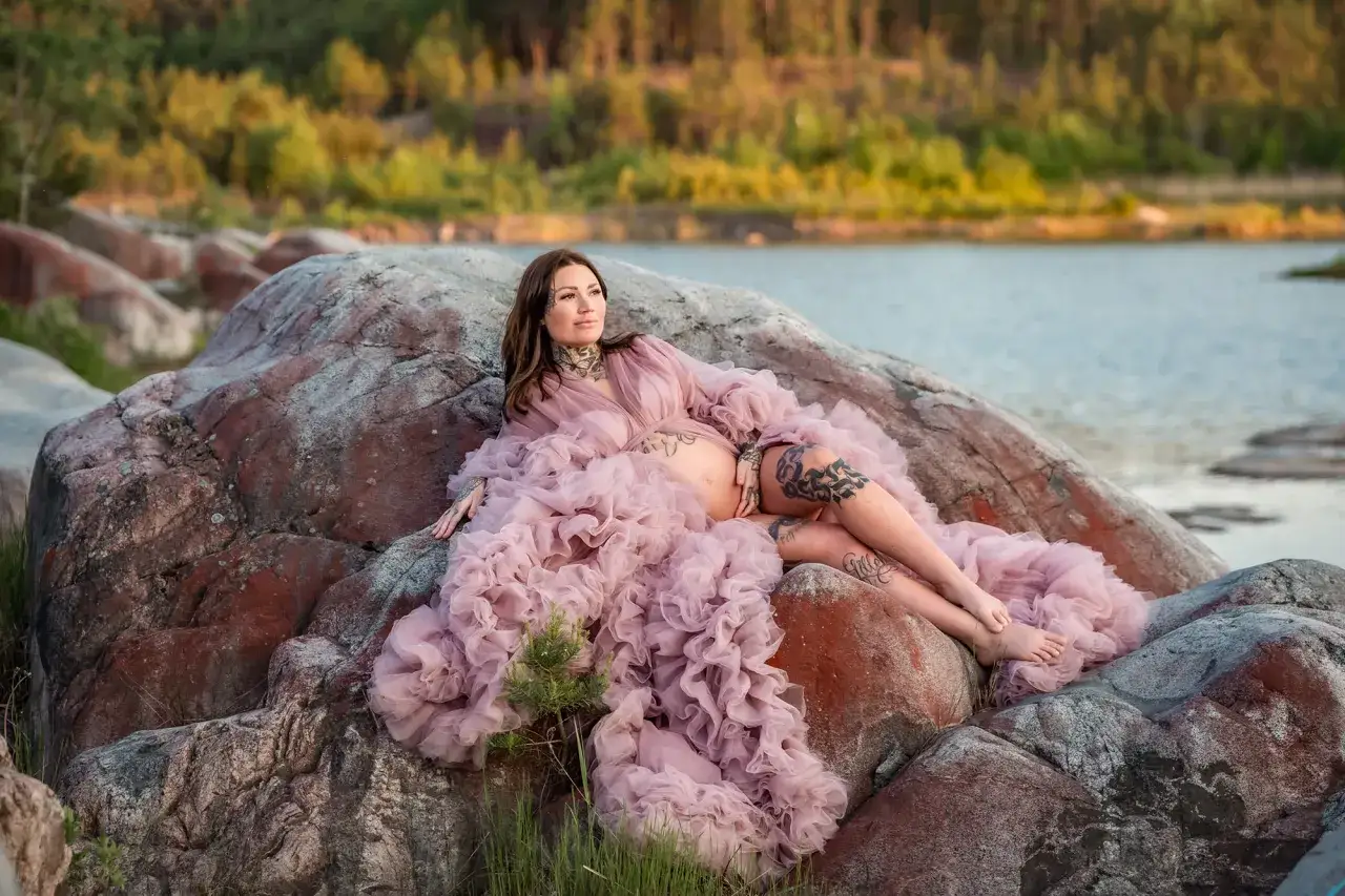En gravid kvinna i en rosa kl&auml;nning vilar p&aring; stenar vid vattnet. Hon njuter av lugnet, en vacker tid att springa gravid.