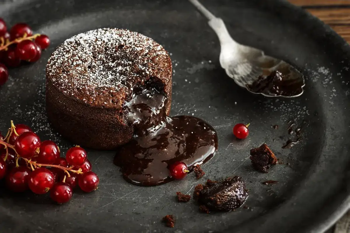 Un delizioso tortino al cioccolato con cuore fuso, cosparso di zucchero a velo, accompagnato da ribes rossi freschi su un piatto scuro.