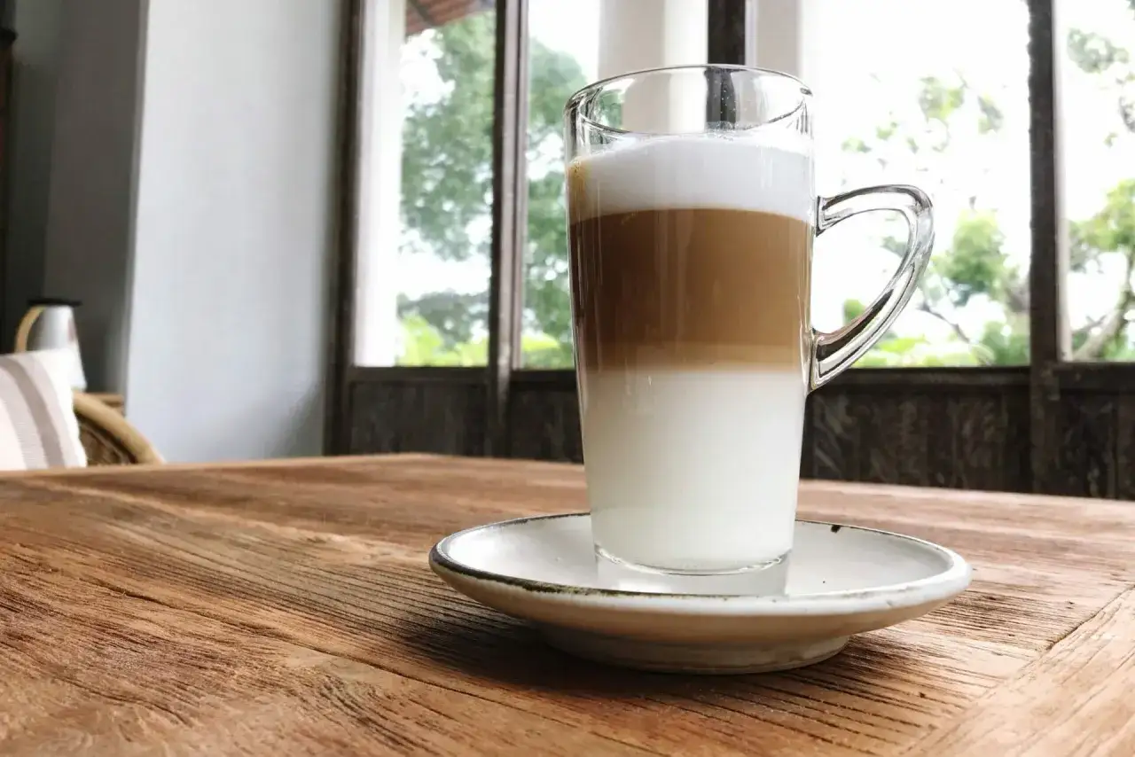 Un caff&egrave; macchiato caldo a strati, servito in un bicchiere trasparente su un piattino bianco, su un tavolo di legno rustico.