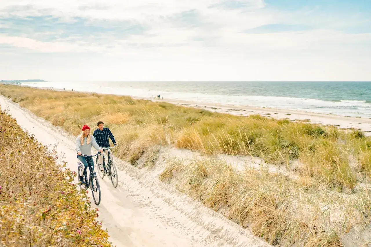 Ein Paar genießt seine baltic adventures auf Fahrrädern entlang eines sandigen Küstenwegs mit Blick auf das Meer und Dünen.