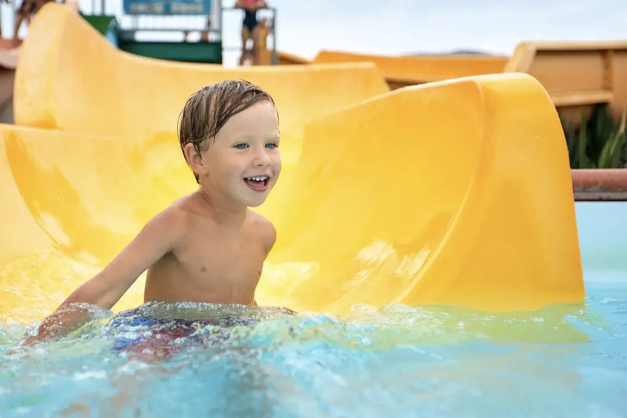 Ein Junge mit blauen Augen lacht fröhlich im Wasser am Ende einer gelben Wasserrutsche im Heidebad Torgelow.