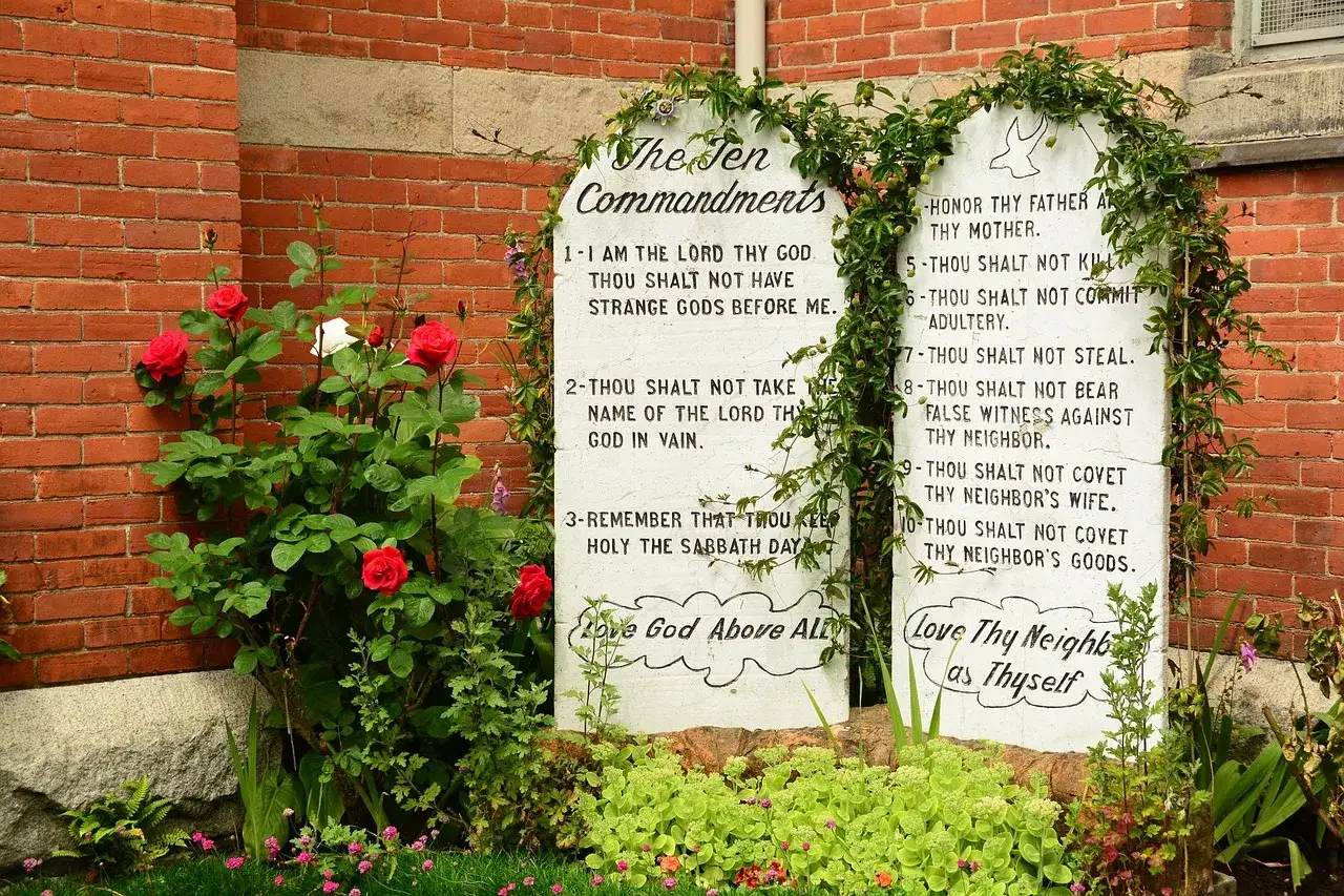 Zwei Steintafeln mit den katholischen 10 Geboten, umrankt von Gr&uuml;nzeug und Rosen, vor einer Ziegelwand.