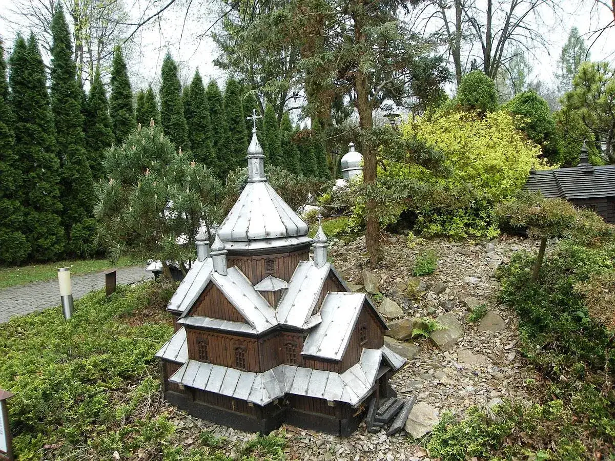 Drewniana cerkiewka z metalowym dachem, miniatura w parku miniatur Myczkowce, otoczona zielenią i kamieniami.