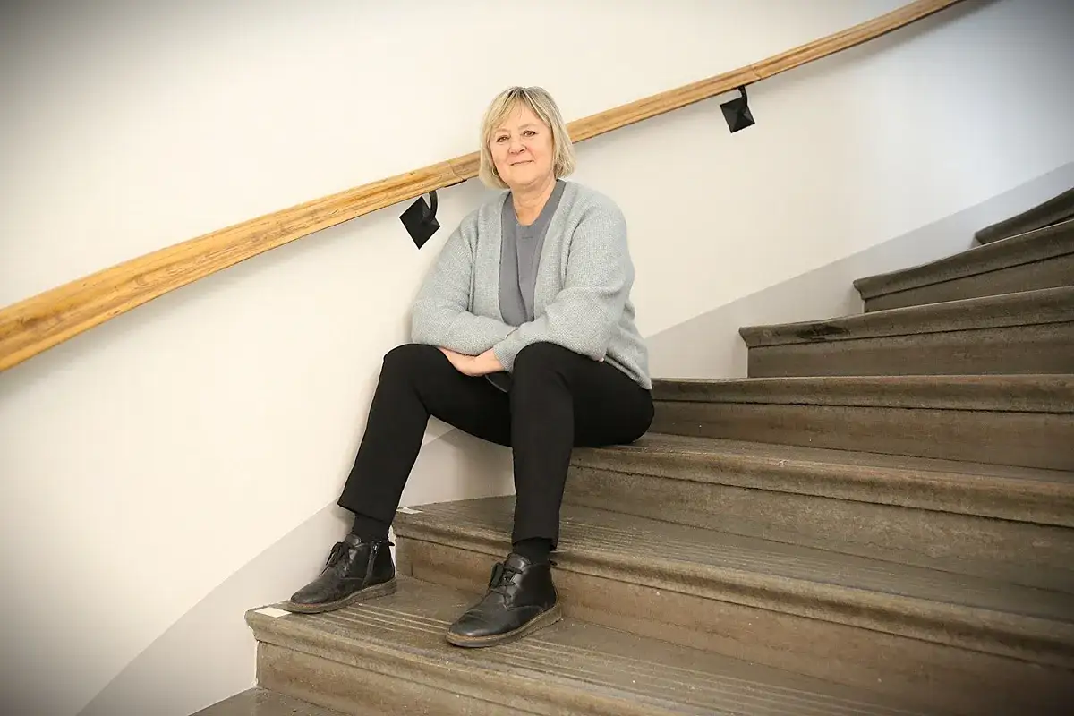Charlotte W&aring;hlin sitter p&aring; en trappa med ett leende. Hon b&auml;r en gr&aring; kofta och svarta byxor.