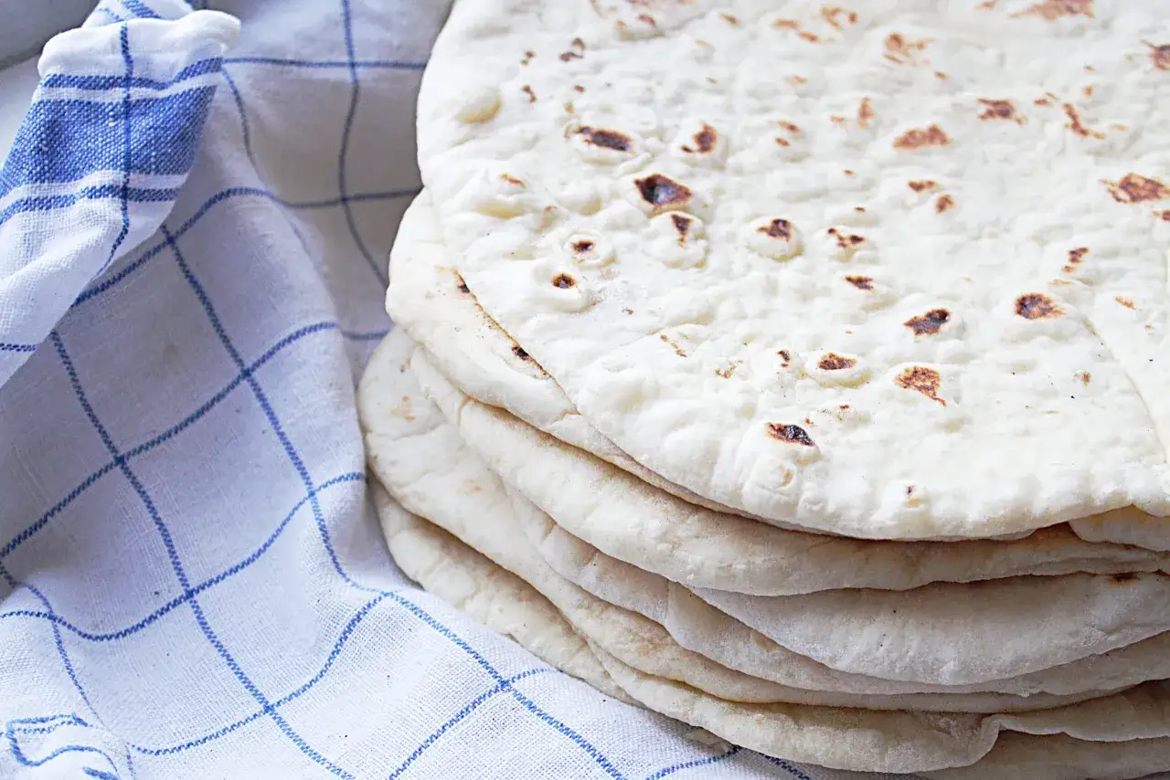En hög med nybakade tortillabröd, perfekta för ditt nästa tortilla recept. Bredvid ligger en blårutig kökshandduk.